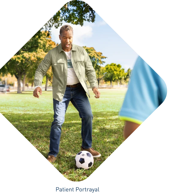 Middle-aged male in park setting preparing to kick soccer ball to second person just out of frame.